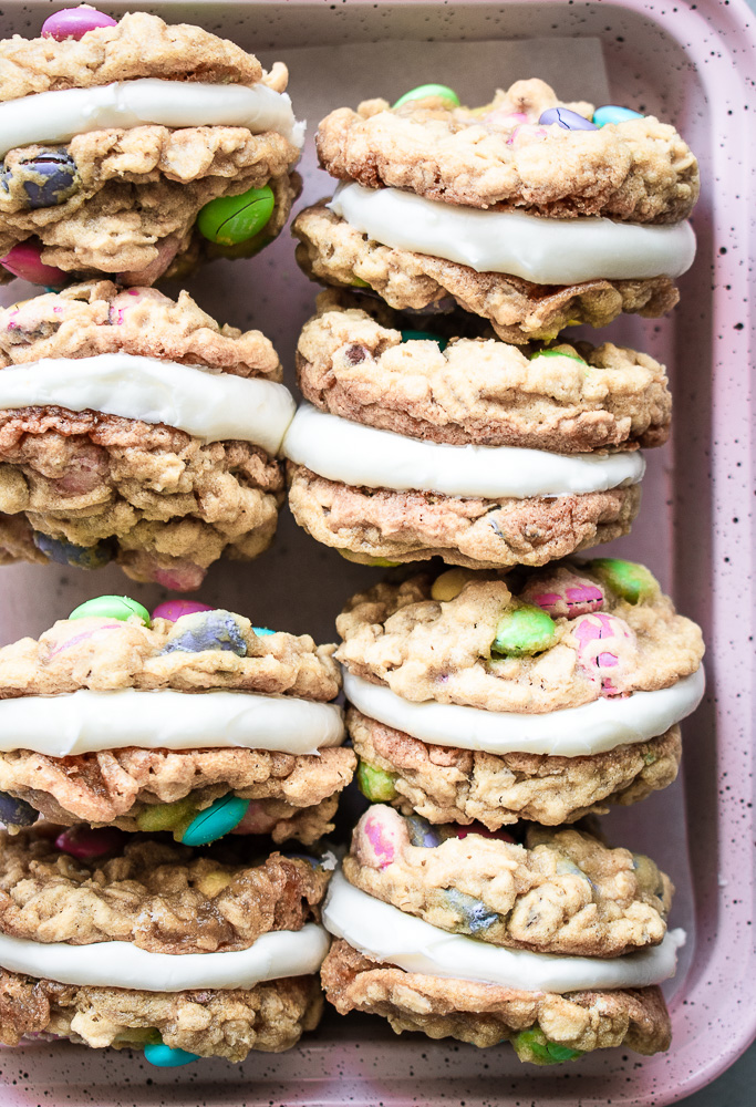 Cream cheese frosting between monster M&M oatmeal cookies on a pink baking sheet.